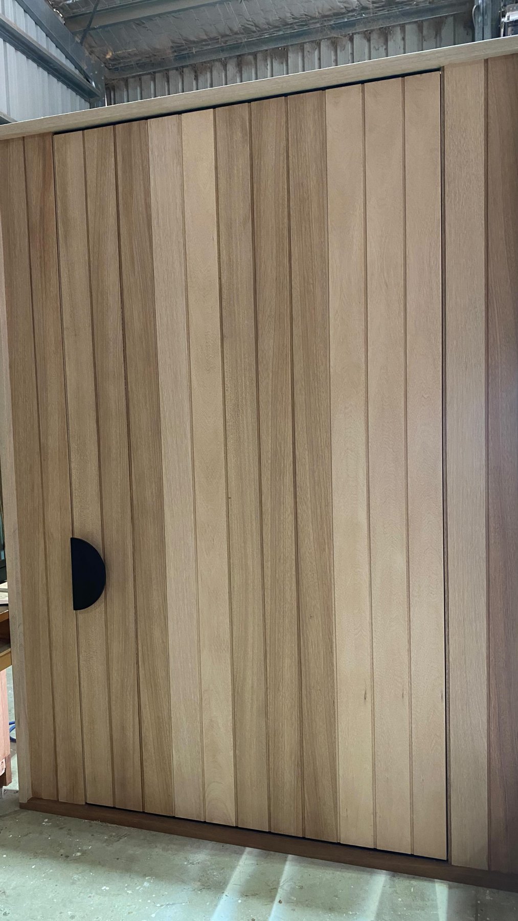 Modern vertical-slat timber entry door with a black half-moon pull handle, workshop closeup